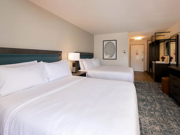 A hotel room with two large beds, white linens, a nightstand lamp, a dresser, a wall art piece, and a seating area near the doorway.