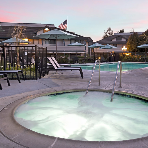 A resort-style pool area at dusk, with a lit hot tub in the foreground, lounge chairs, umbrellas, and a clubhouse in the background.