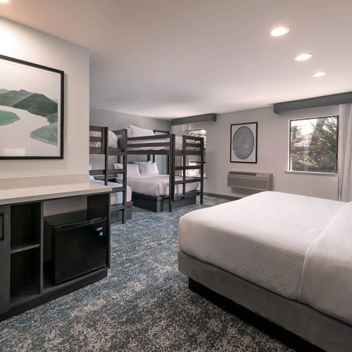 A spacious hotel room with two queen beds, a bunk bed area, kitchenette, workspace, and large window; modern decor and soft lighting.