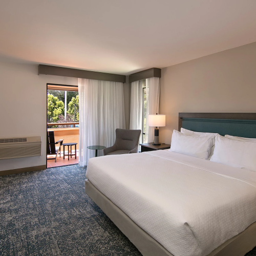 A modern hotel room with a queen bed, blue-grey headboard, two nightstands, lamps, a seating area by the window, and a balcony visible outside.
