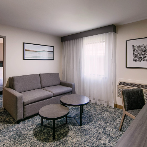 A modern hotel suite with a gray sofa, two round coffee tables, a desk and chair, framed art, a large window with sheer curtains, and a doorway to a bedroom.