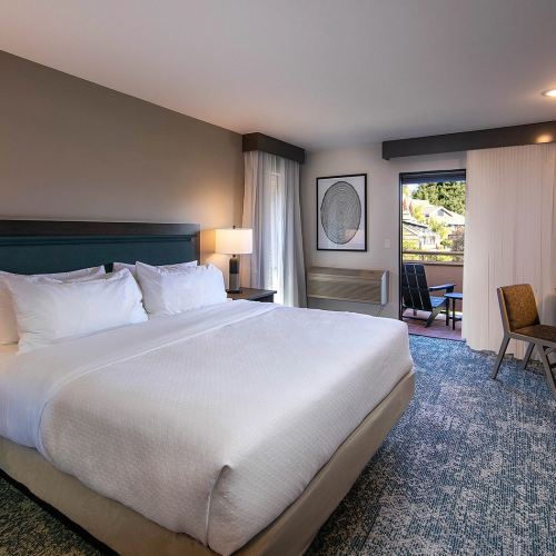 A modern hotel room with a king bed, nightstands and lamps, a flat-screen TV, a desk, and a balcony door letting in natural light.