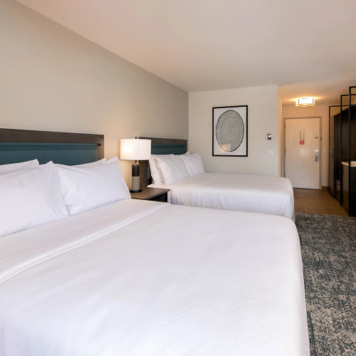 A hotel room with two large beds, white linens, a nightstand and lamp between them, a dresser, artwork, and a doorway leading to a small bathroom; bright, clean, modern.