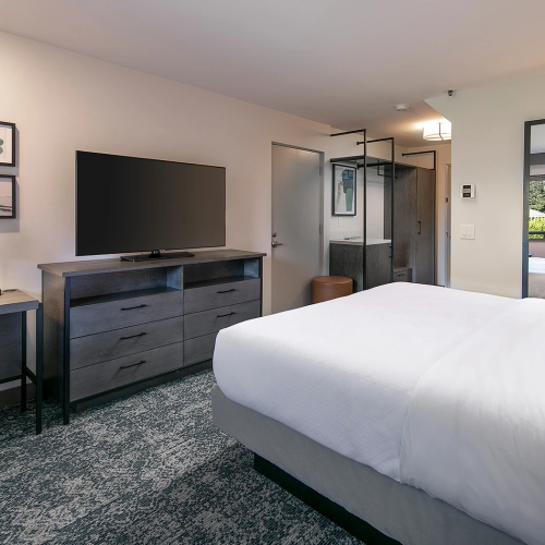 A modern hotel room with a large bed, desk and chair, flat-screen TV on a dresser, framed art, and a glass-enclosed bathroom in the background.