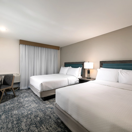 Two double beds in a modern hotel room with a desk, chair, flat-screen, and a nightstand between beds; neutral tones and a blue accent.