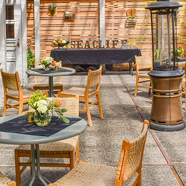 Outdoor patio with wooden chairs, round tables, potted plants, a tall heater, and a banner that says &ldquo;SAG??&rdquo; under a wooden screen.
