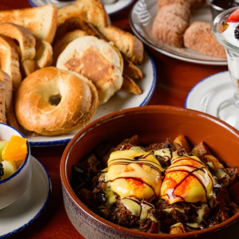 A spread of breakfast foods: bagels and bread, a fruit parfait, a yogurt parfait, a bowl of bubbling chocolate and ice cream with drizzle, and fresh fruit on the side.