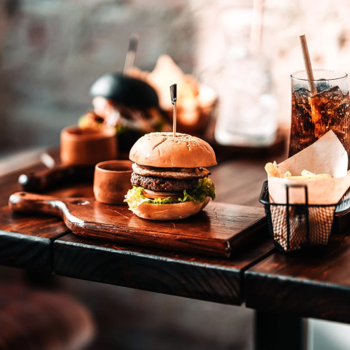A wooden table with a burger, fries in a basket, and a glass of soda; a second burger and drink sit in the background on a rustic setting.