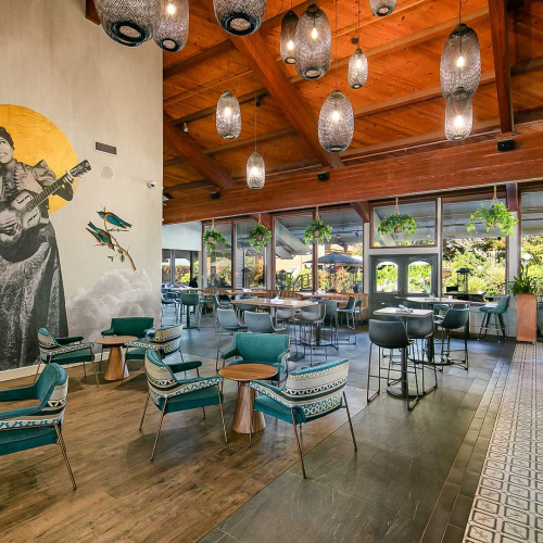 A stylish, sunlit lounge with teal chairs, wooden tables, hanging lanterns, a mural of a guitar player, and large windows overlooking greenery.