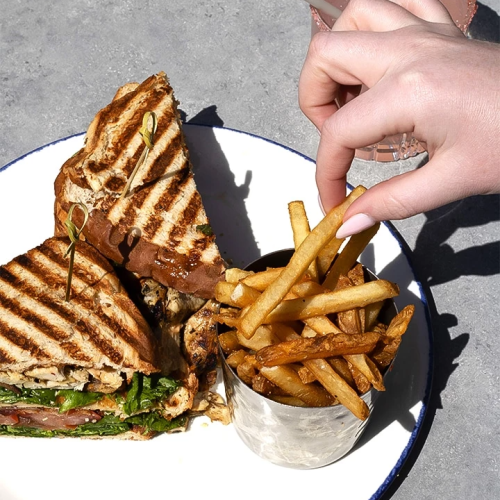 A plated grilled sandwich with melted greens, a side of fries, and a hand dipping a drink cup on a gray surface.