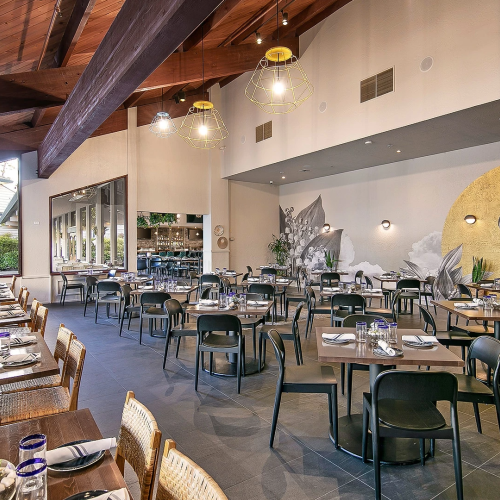 A bright, modern dining room with wooden beams, many tables set with plates and glassware, stylish pendant lights, and wall art featuring a pelican mural.