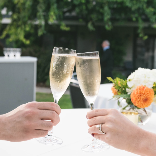 Two people clinking champagne flutes at a table outdoors with a floral centerpiece in the background, celebrating together.