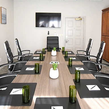 A modern conference room with a long table, black leather chairs, notepads and green glass cups, a wall-mounted TV, cabinets, and plants.