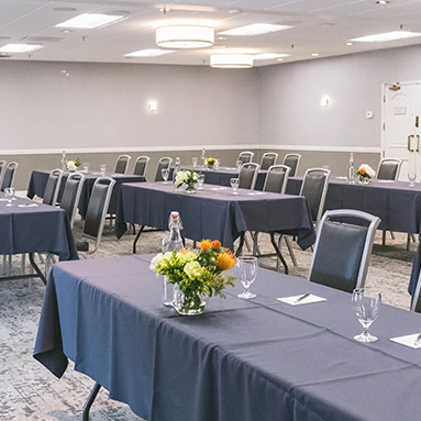 A conference room set up for a meeting with long navy tables, flowers, glasses, and chairs arranged in rows.