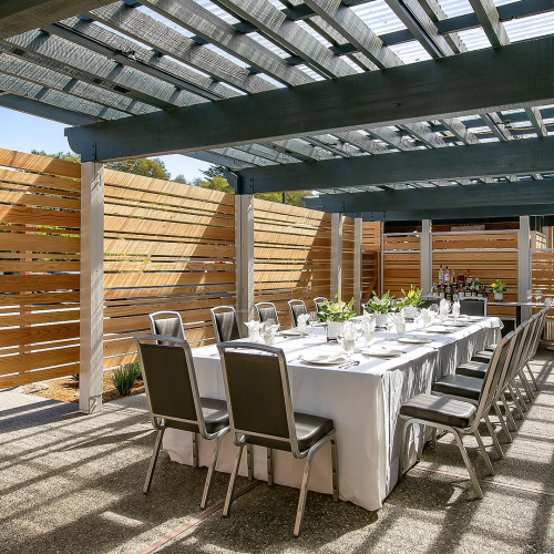 A long dining table set for a meal under a covered outdoor patio with wooden slat walls and ample natural light.