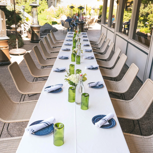 A long outdoor dining table set for a meal, with white tablecloths, neatly arranged place settings, green glass bottles, and wicker chairs under bright daylight.