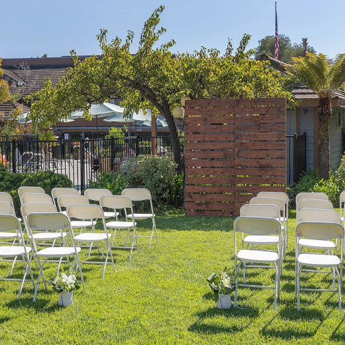 A sunny outdoor wedding setup with white folding chairs arranged in rows on a green lawn, a wooden arch/trellis, and decorative plants.