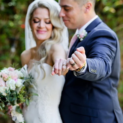 A newly married couple pose outdoors, showing off rings; the bride smiles with a bouquet, while the groom in a navy suit highlights their rings.