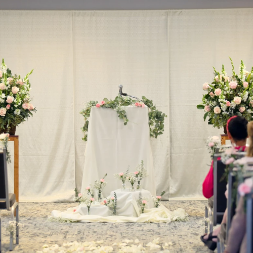 A wedding altar with a white draped backdrop, floral arrangements, and guests seated on both sides in a softly lit ceremony space.
