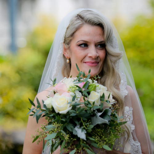 A bride in a veil holds a lush bouquet of white and pink roses and greenery, smiling softly in a garden setting.