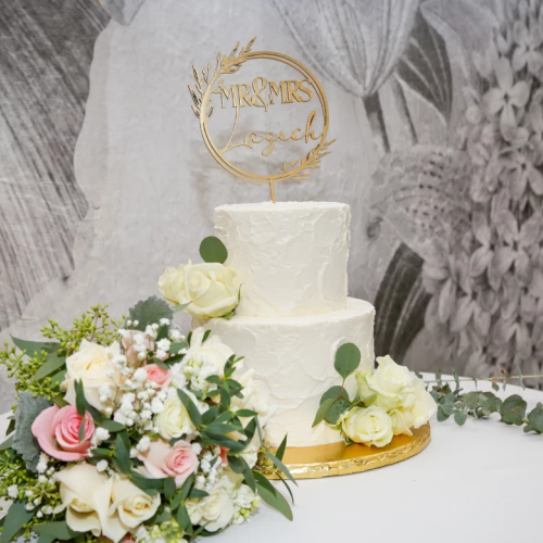 A two-tier white wedding cake with delicate piping, gold base, and a floral bouquet of pink and cream roses, greenery, on a round table with backdrop.