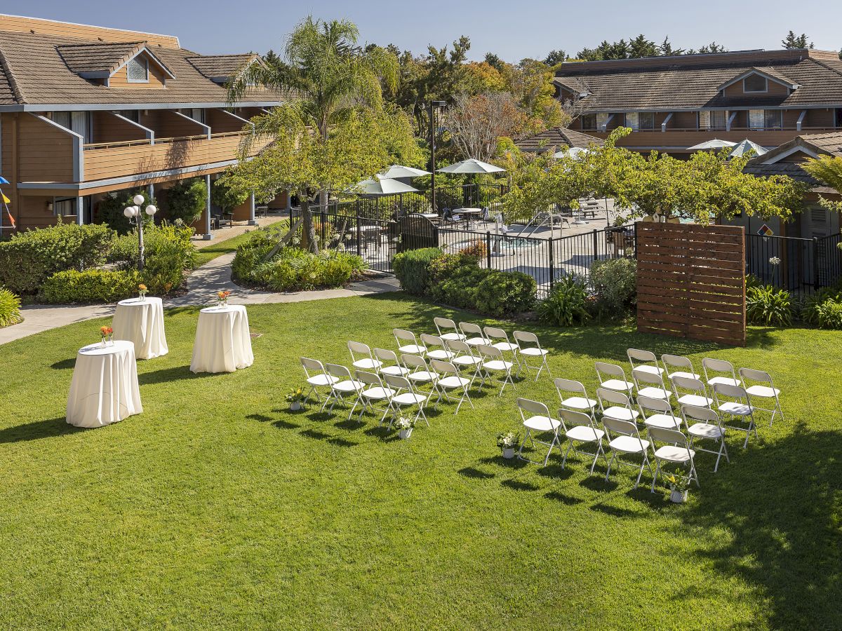 An outdoor garden wedding setup with rows of chairs, round tables, umbrellas, lush landscaping, and surrounding buildings under a clear sky.