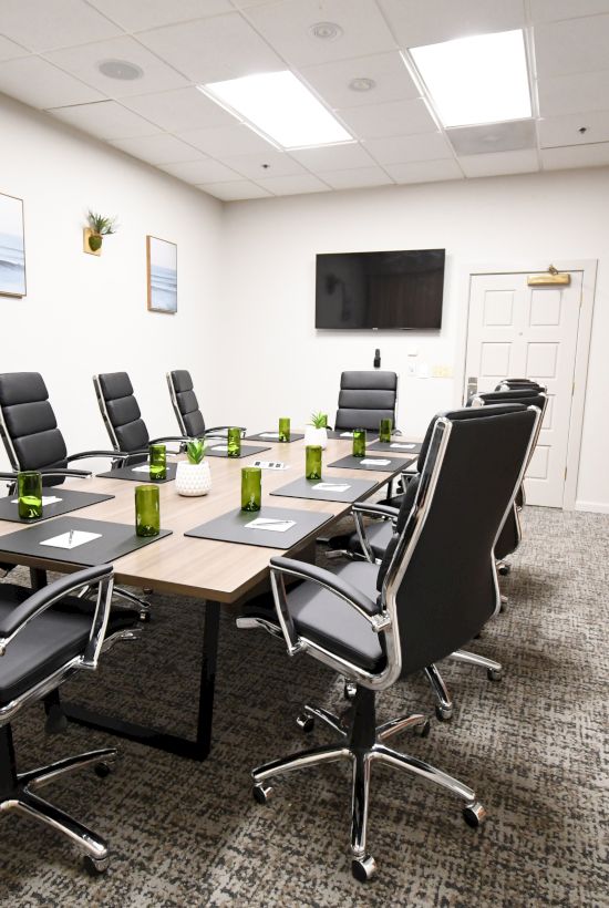 A modern meeting room with a long table, black swivel chairs, green decor, wall TV, and wood cabinets.