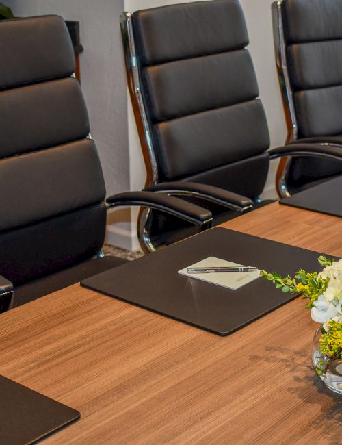A modern meeting room with black leather chairs, a wooden conference table with notebooks, and a vase of white flowers.